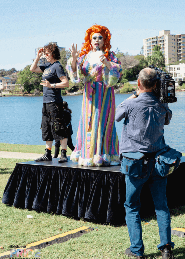 A performer in a rainbow bodysuit poses on a small riverside stage while a photographer works nearby.