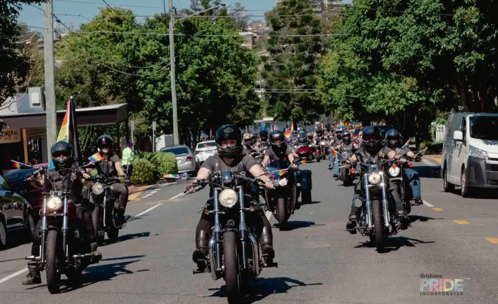 A line of motorcycle riders leads the Pride March down a tree-lined street.