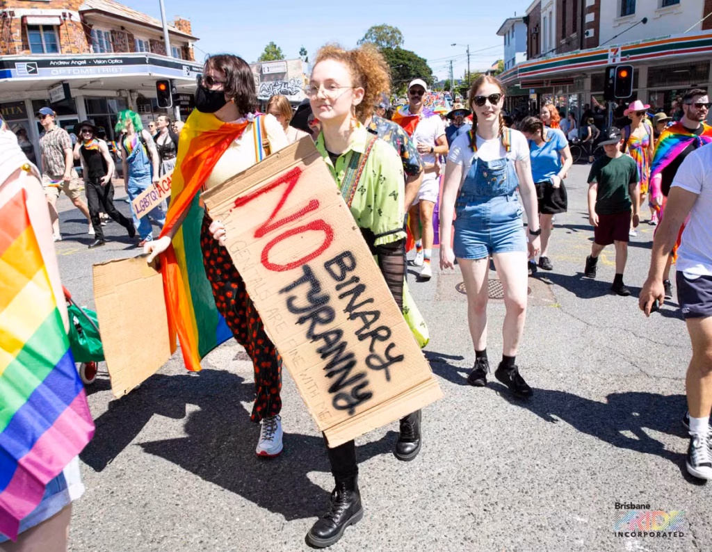 A marcher carries a hand-lettered cardboard placard and a rainbow flag along the march route.