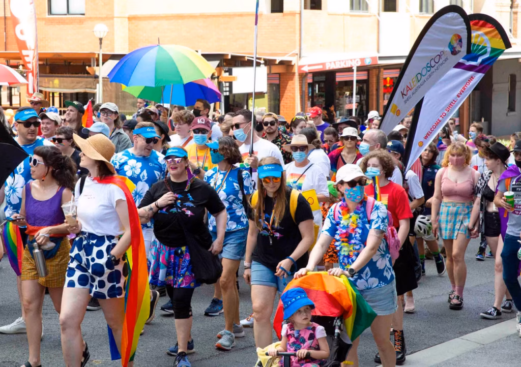 Marchers fill the street under a rainbow umbrella, many in matching shirts, with flags flying above the crowd.