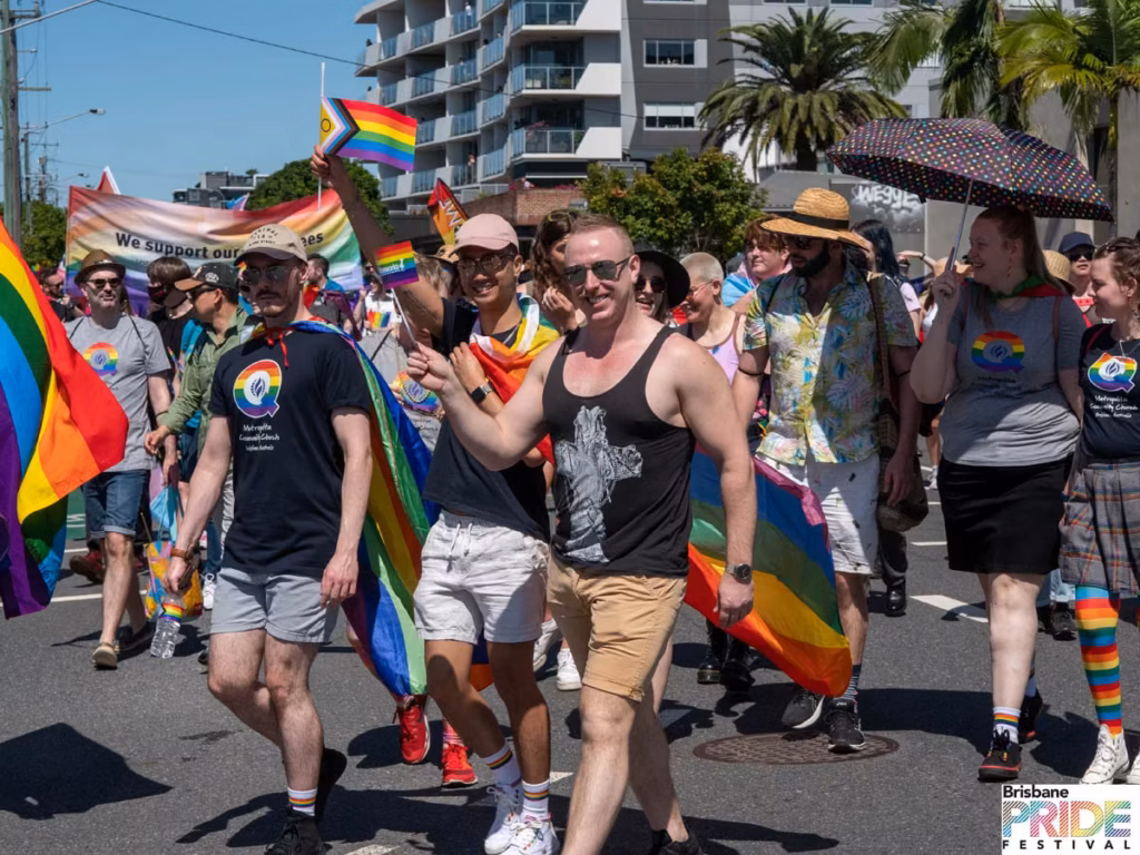 Marchers carrying rainbow flags along a street lined with apartment buildings and palm trees.