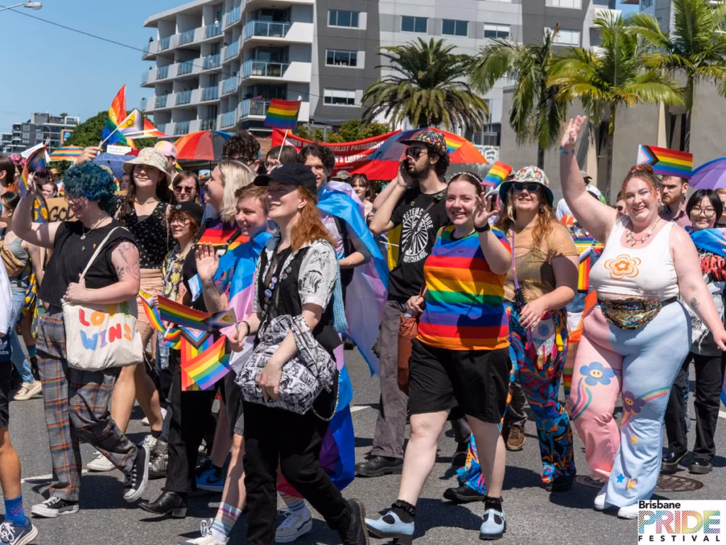 A large group of marchers in rainbow clothing filling a street, with an apartment block and palm trees behind.