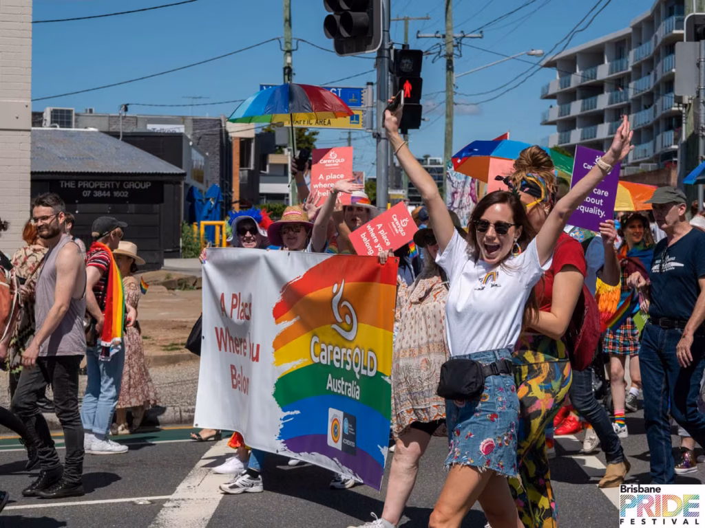 Marchers behind a Carers Queensland rainbow banner, one with arms raised in celebration, at a street intersection.
