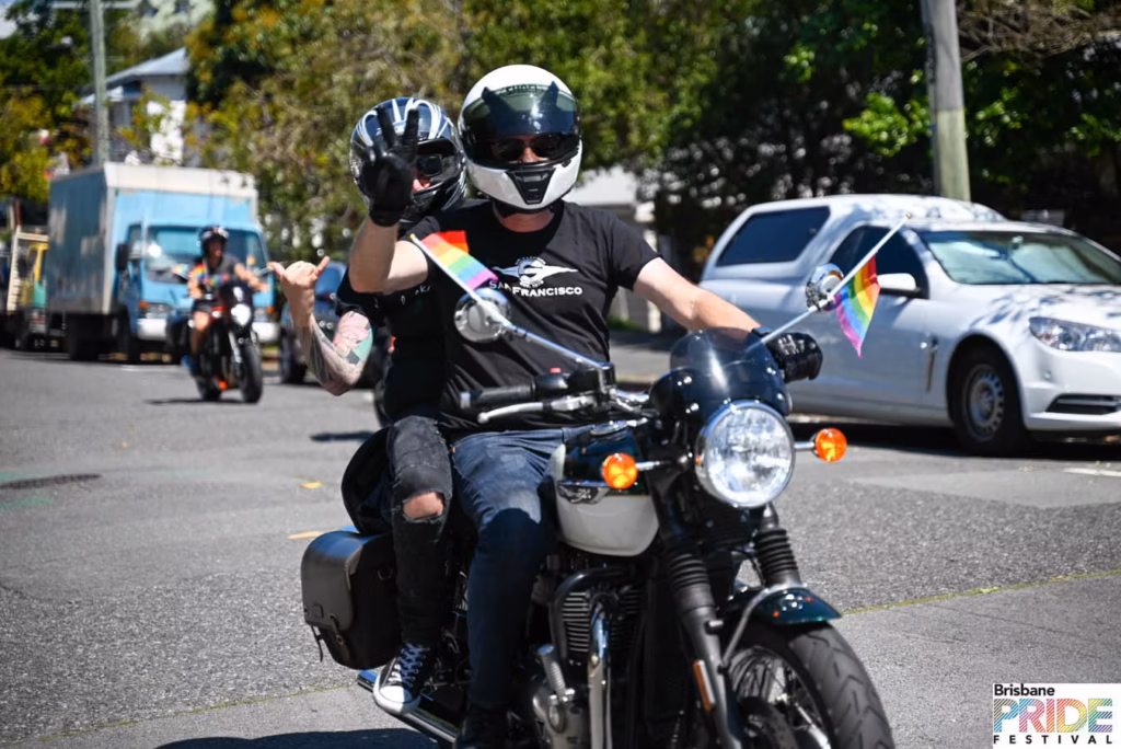 Two riders in helmets on a motorcycle leading the Pride March, small rainbow flags mounted on the bike, one rider waving.