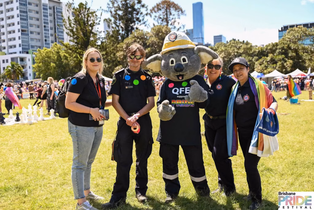 Queensland Fire and Emergency Services staff and a koala mascot posing on the grass at New Farm Park, with the Brisbane city skyline behind.