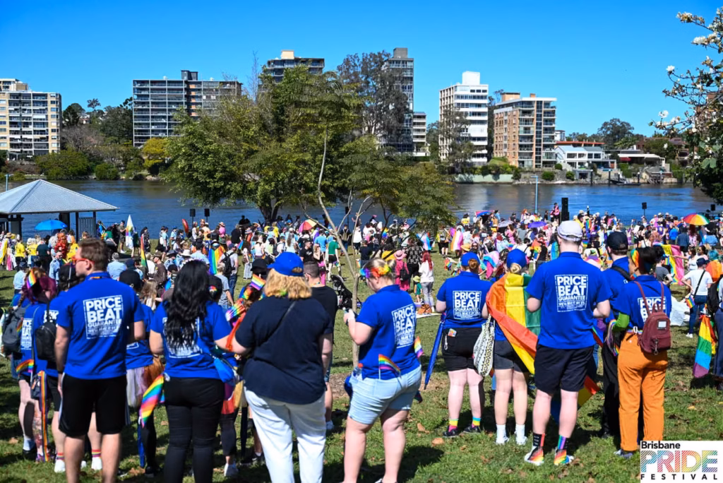 A large crowd on the grass at New Farm Park beside the Brisbane River, many wearing blue Pride Beat t-shirts and carrying rainbow flags, with apartment buildings across the water.