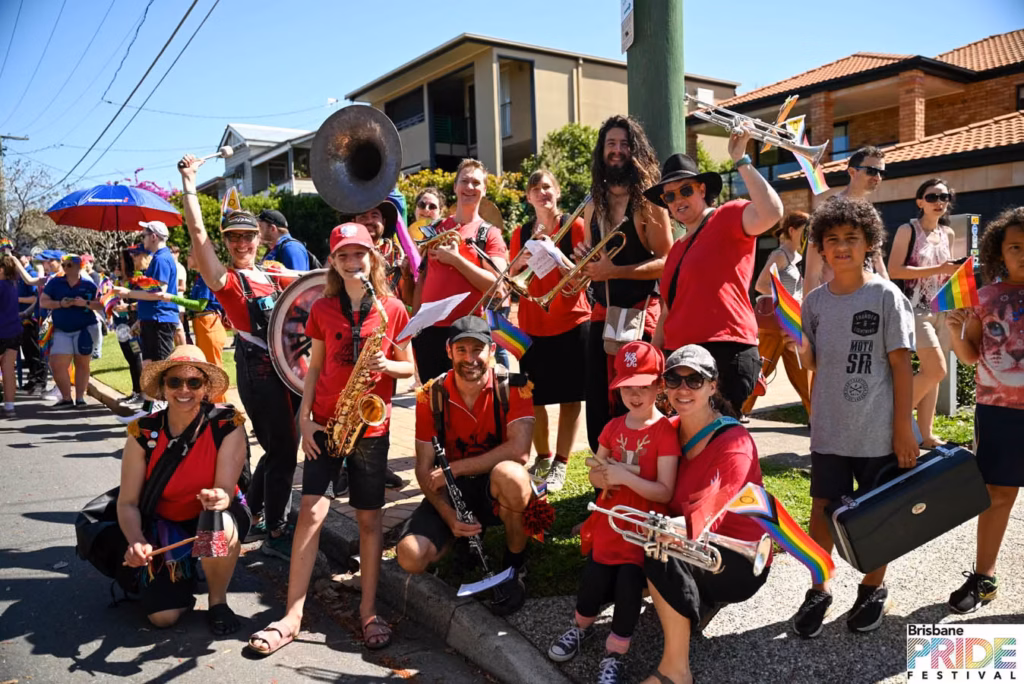 A marching band in red shirts with tuba, saxophones and trumpets gathered on a suburban street during the Pride March.
