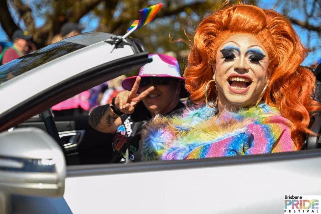 A drag performer in a bright orange wig and rainbow fur stole leaning out of a white convertible during the Pride March, a rainbow flag mounted on the car.