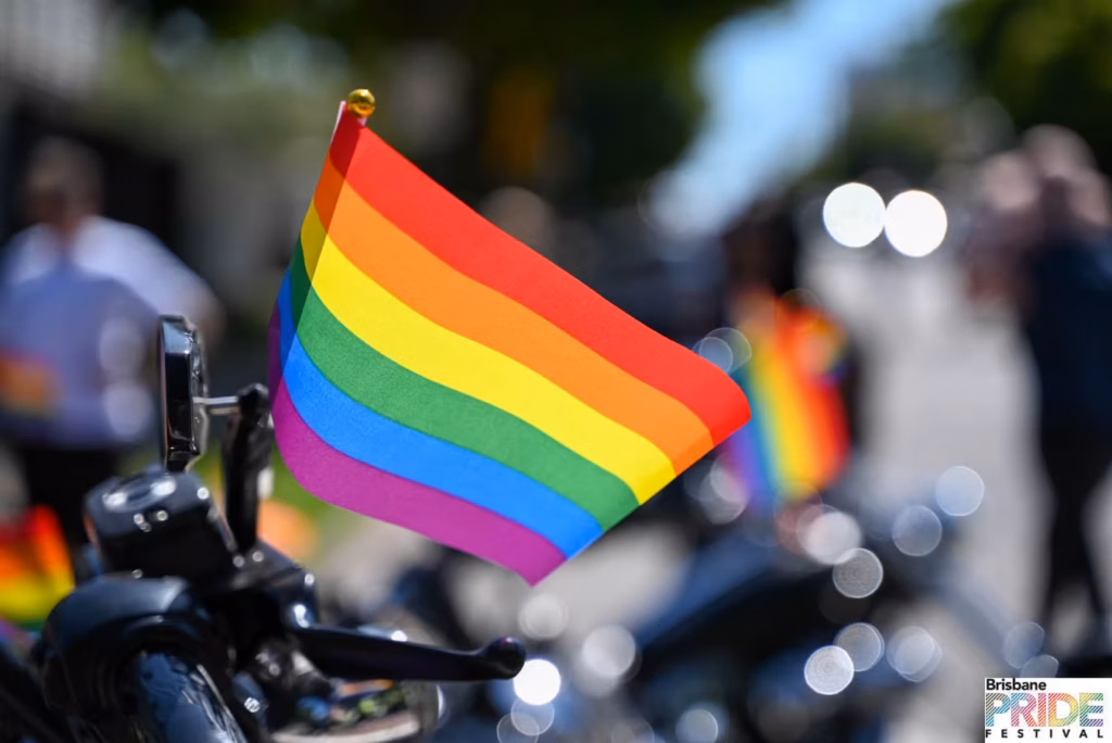 A rainbow flag mounted on a motorcycle handlebar, with a blurred crowd behind.