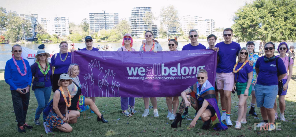 A workplace group gathers on the grass behind a purple we all belong banner, with the river and the city behind them.